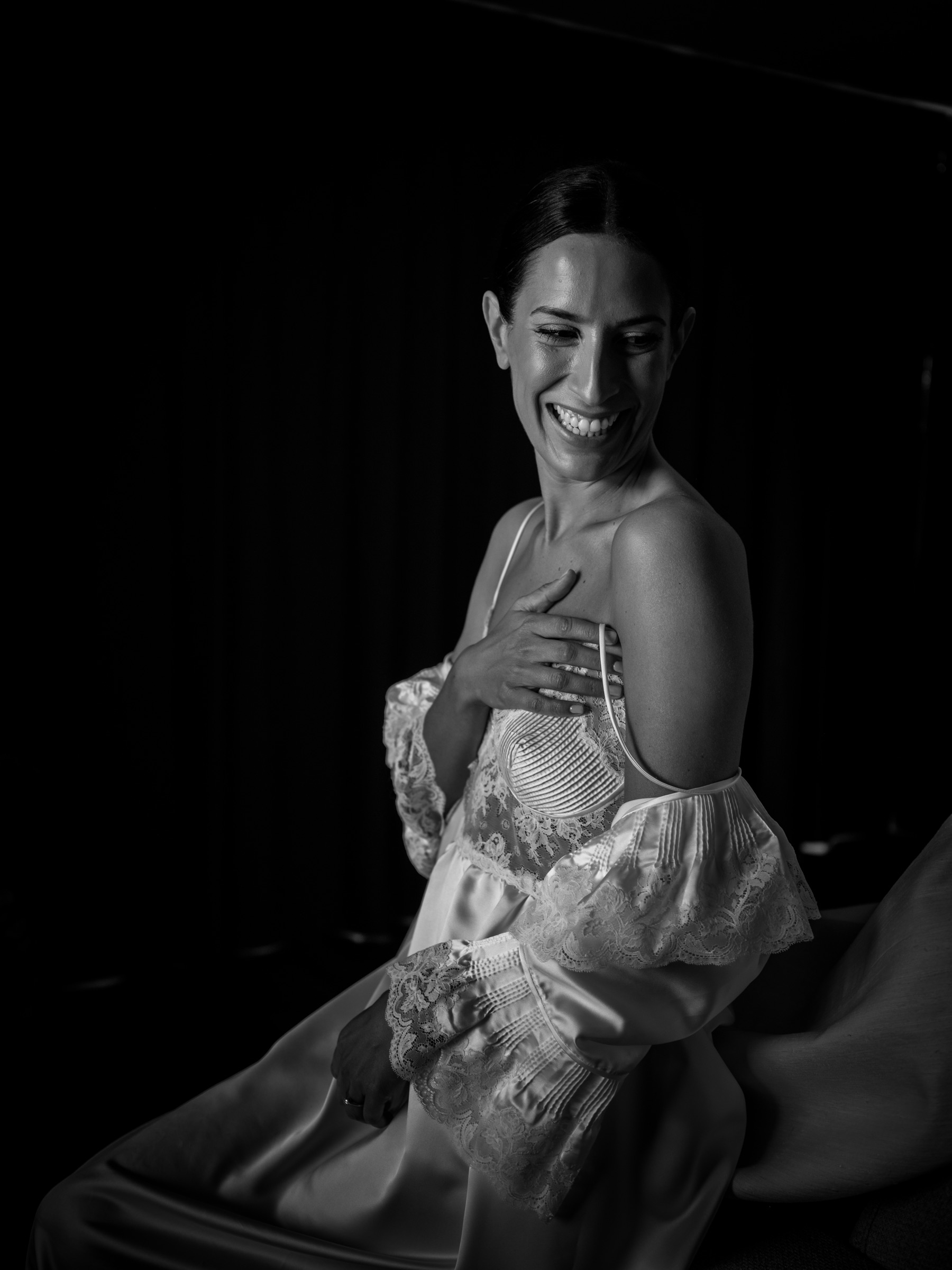 Bride seated in a lace-edged silk slip, laughing with a hand to her chest, carved out of the darkness of the room by a single window