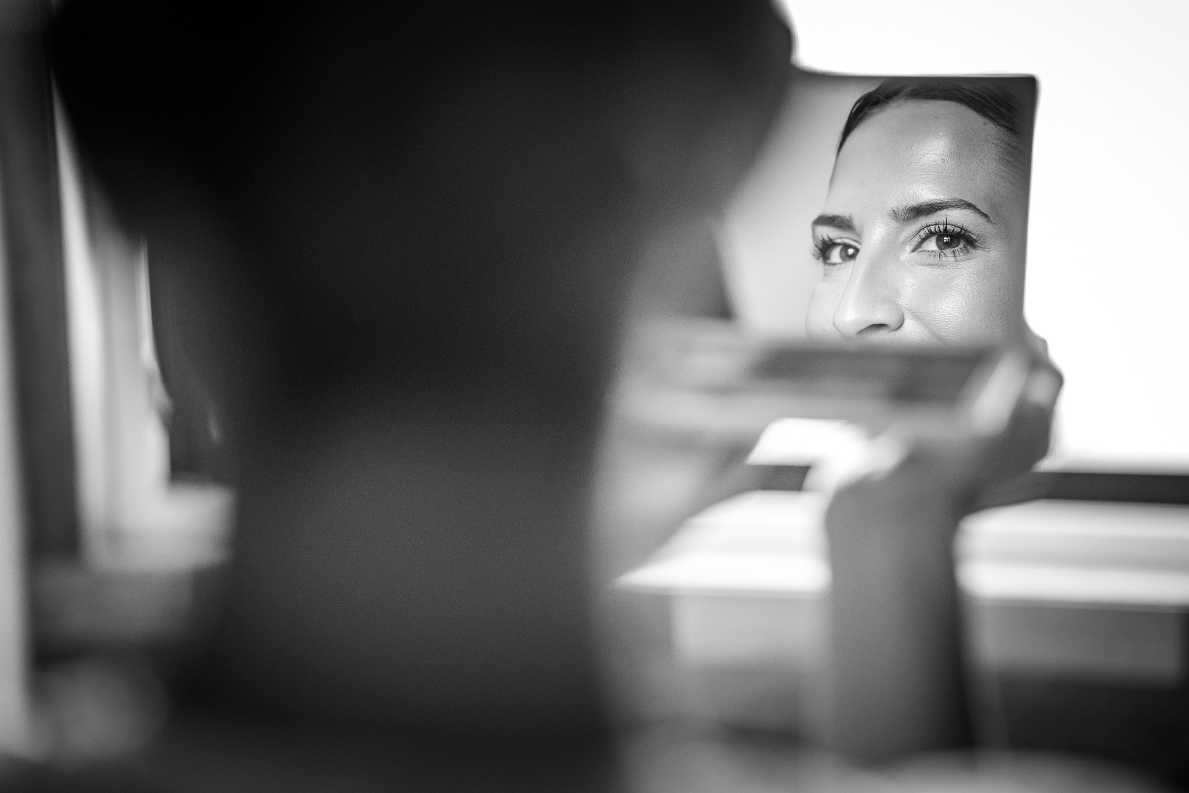 The bride's made-up eyes caught in a hand mirror, the makeup artist a soft dark silhouette in the foreground