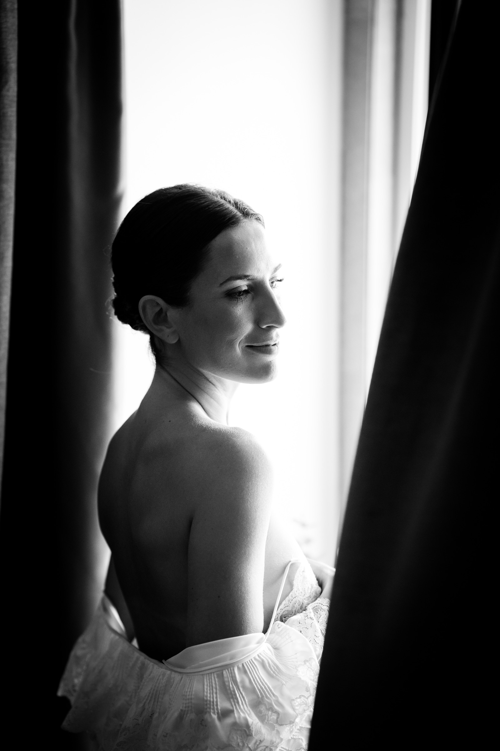 Bride glancing back over her bare shoulder between dark curtains, holding her dress to her chest in the window light
