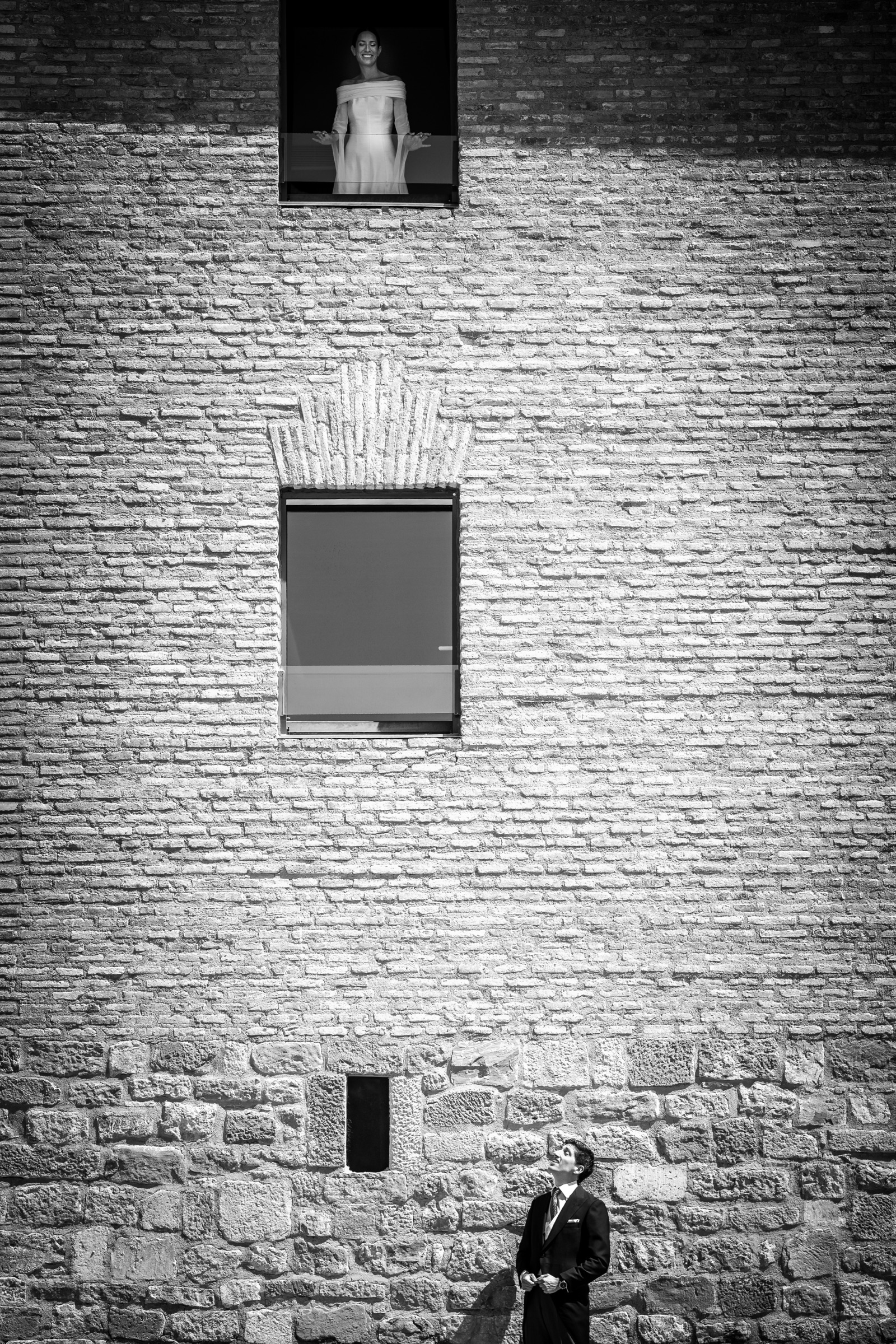 Bride opening her arms at the top window of the monastery's vast brick wall while the groom looks up from the street below