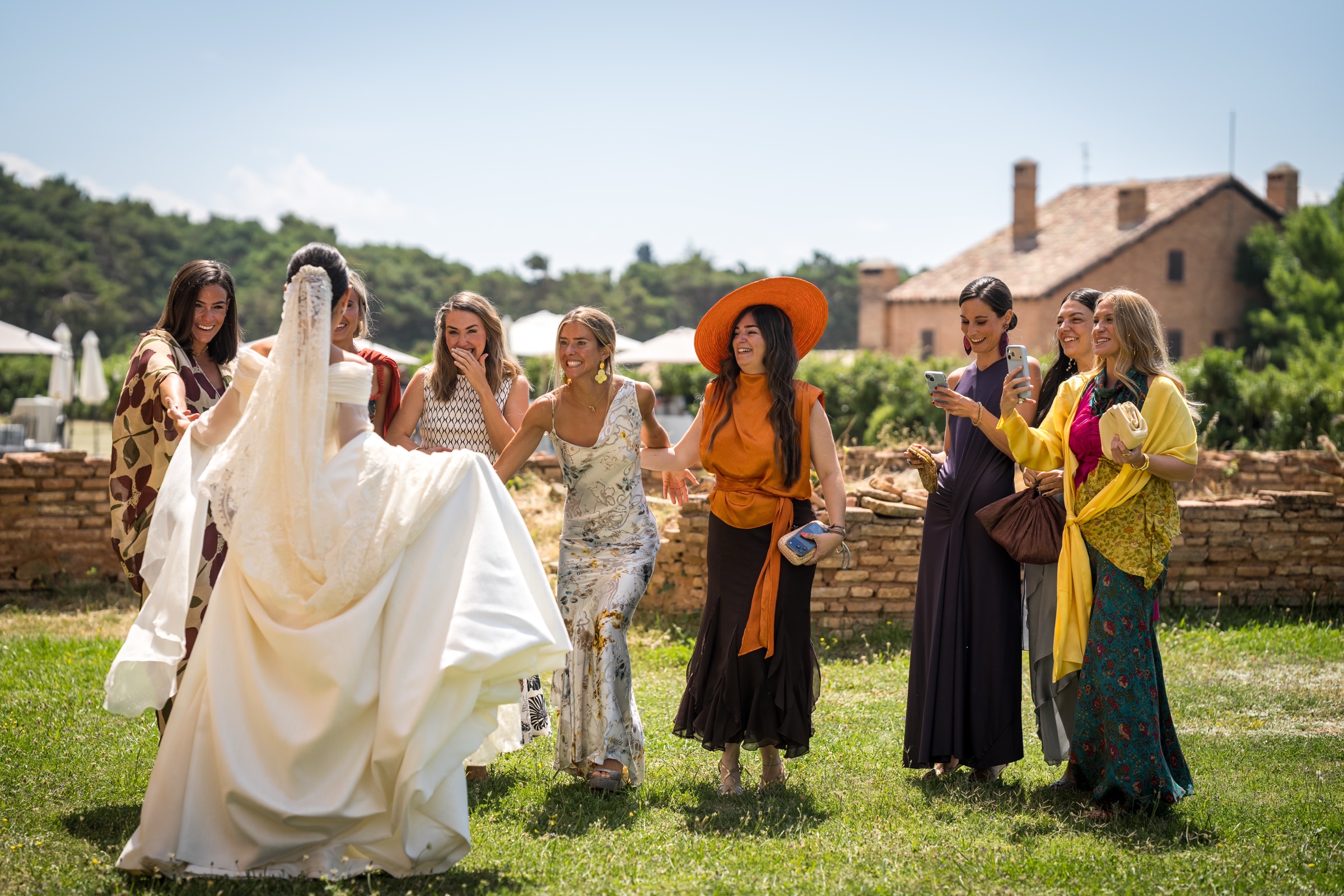 Friends in summer colour crowd laughing around the bride's lace mantilla veil on the sunlit lawn