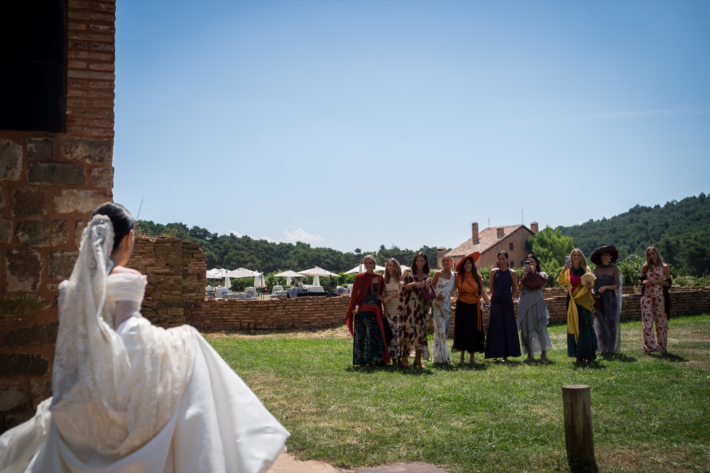 Seen past a brick corner, the bride crosses the meadow above San Juan de la Peña toward a waiting line of friends under a vast midday sky