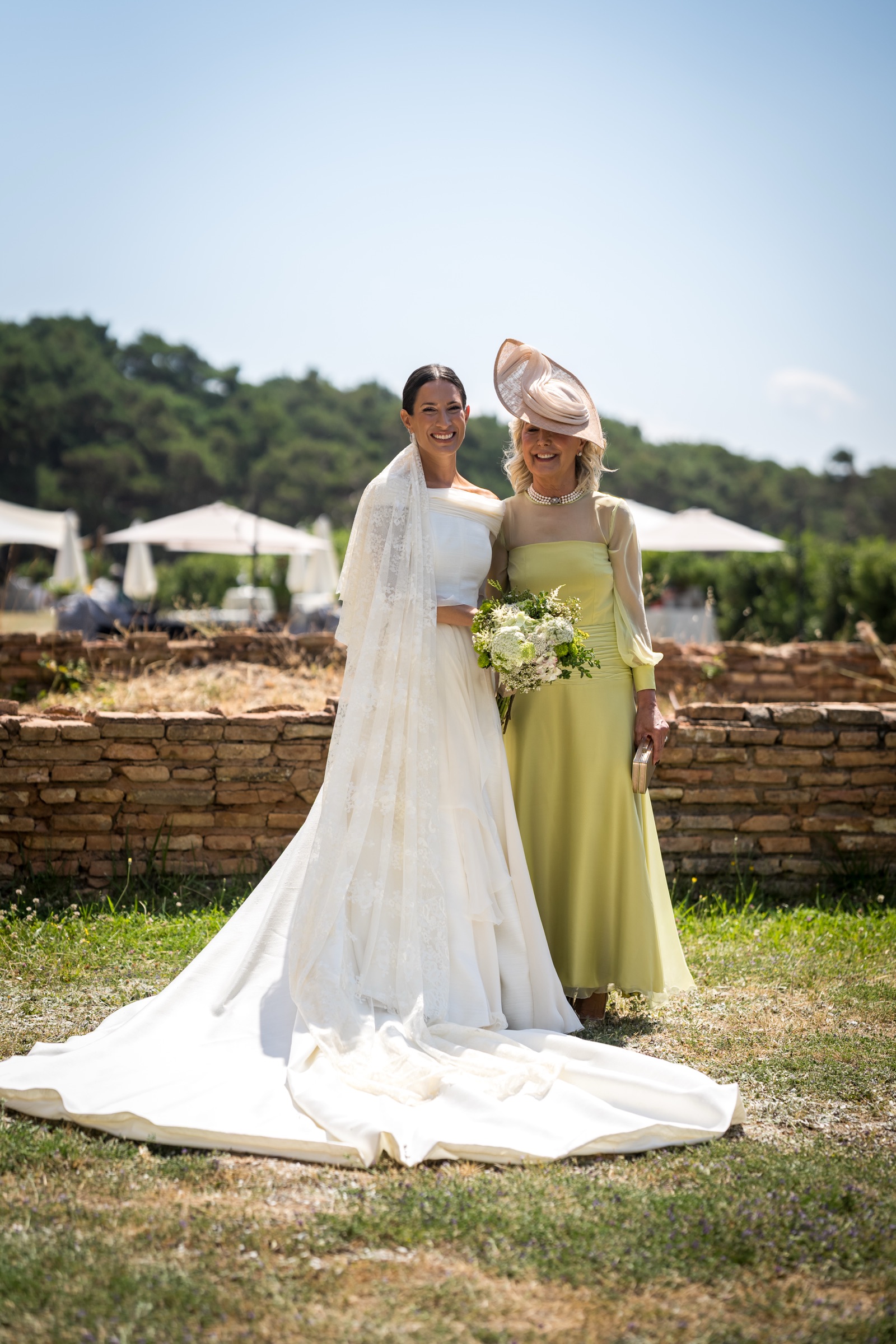 The bride and her mother in pistachio green arm in arm before a low brick wall and white parasols, a loose bouquet between them