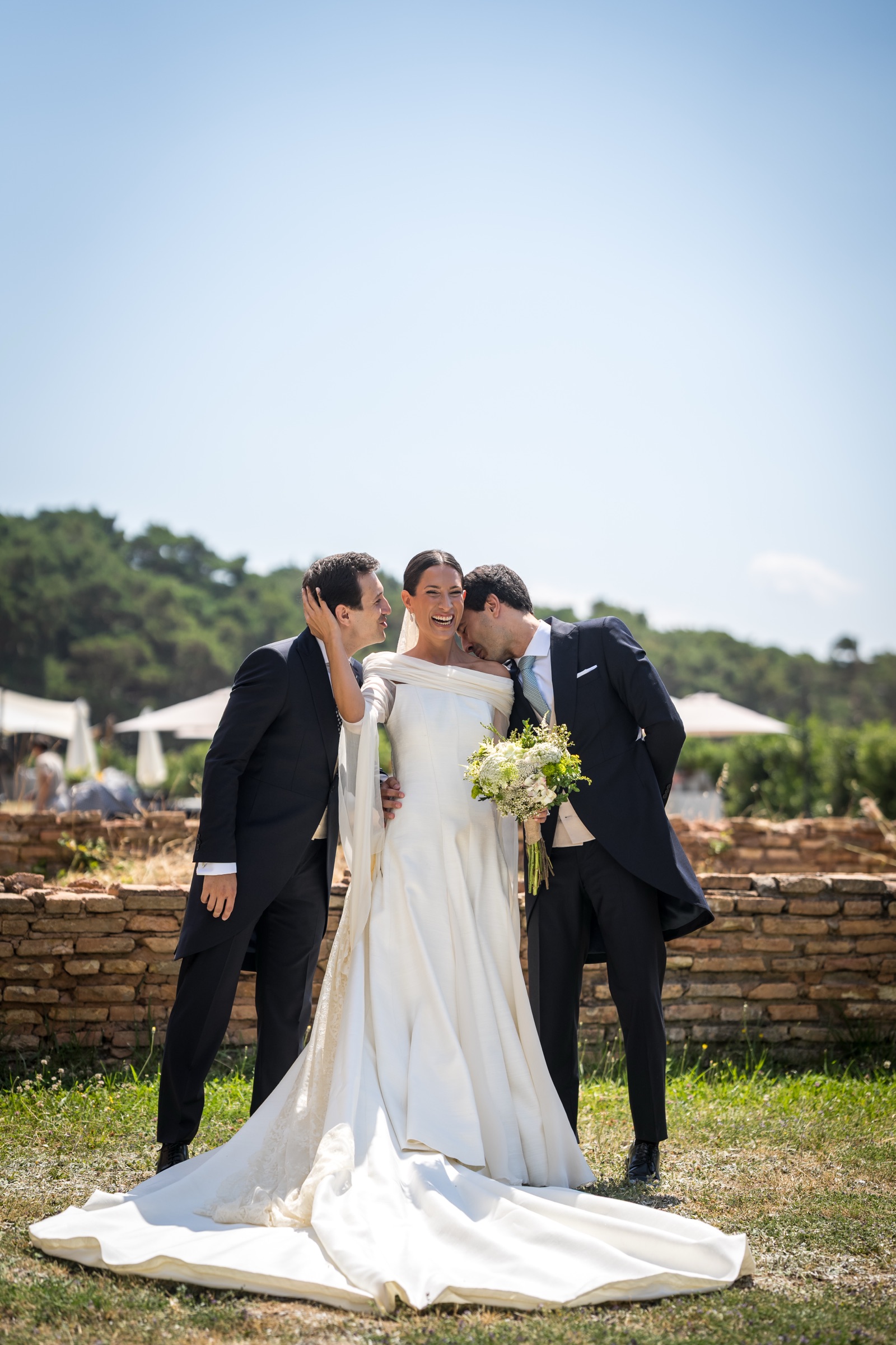 Two men in morning coats kiss the bride's cheeks at once as she laughs, her satin train pooling on the grass