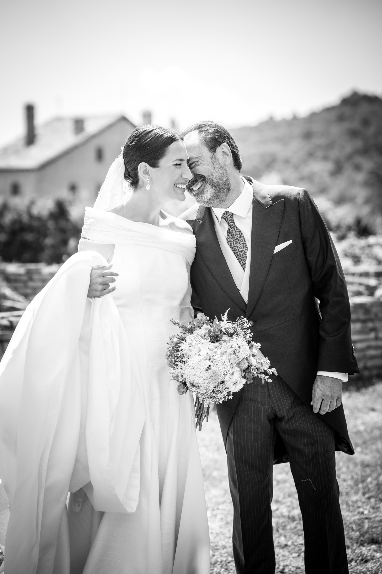 Nose to nose with her father in his pinstriped morning suit, the bride laughs into the backlight, a wildflower bouquet caught between them