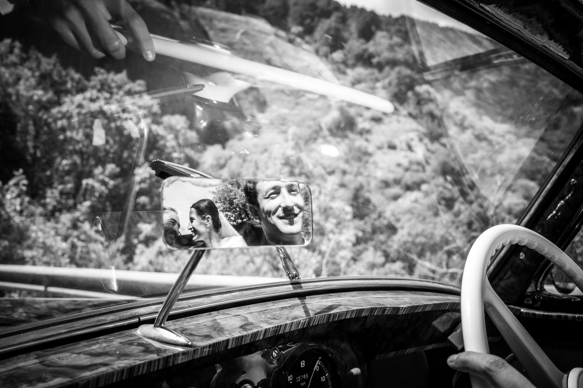 The bride and a grinning driver caught in the rear-view mirror of a vintage car, the wooded mountainside of San Juan de la Peña beyond the windshield