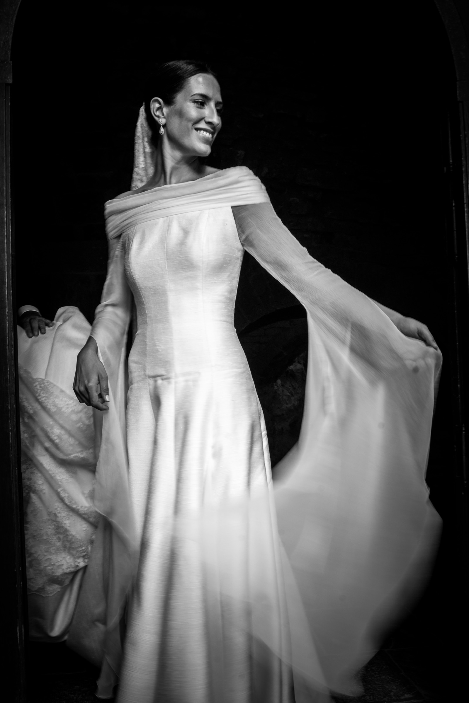 Bride turning in a shadowed monastery archway, chiffon sleeves fanned wide as her silk skirt catches the light