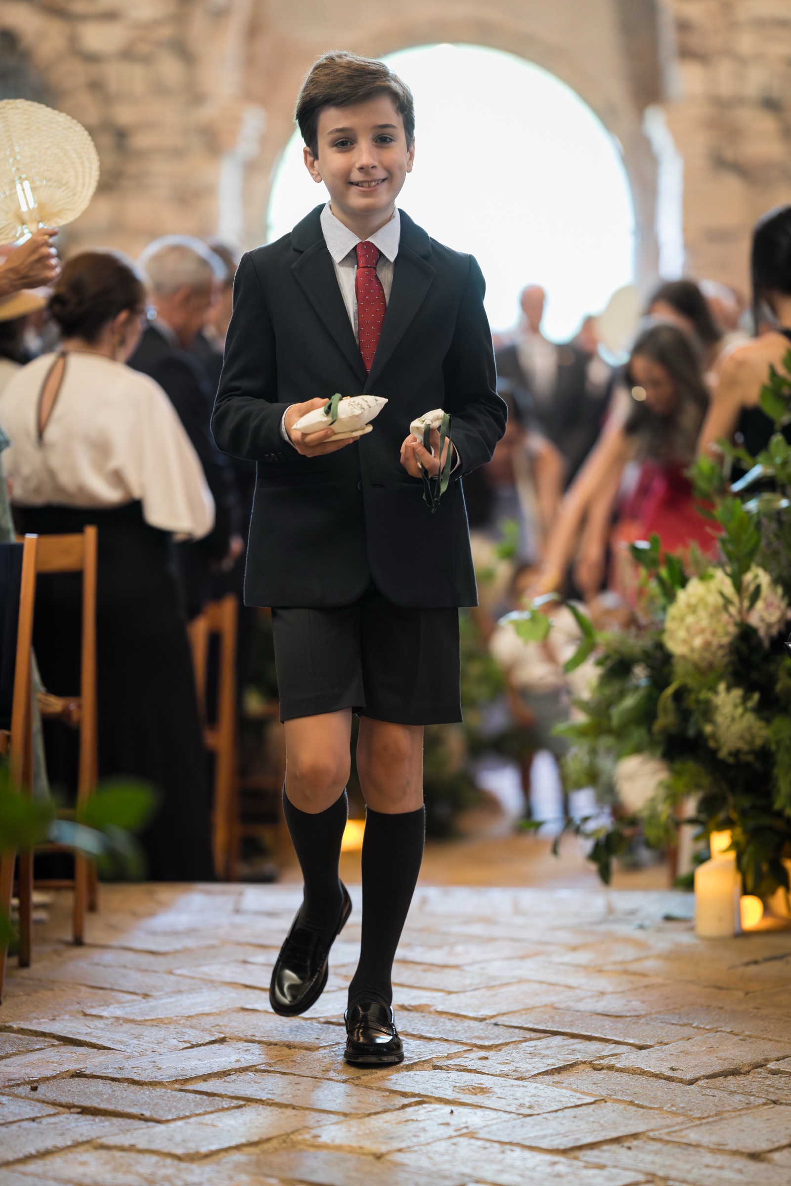 A page boy in shorts and knee socks carrying the ring cushions down the brick aisle beneath the monastery's stone arch