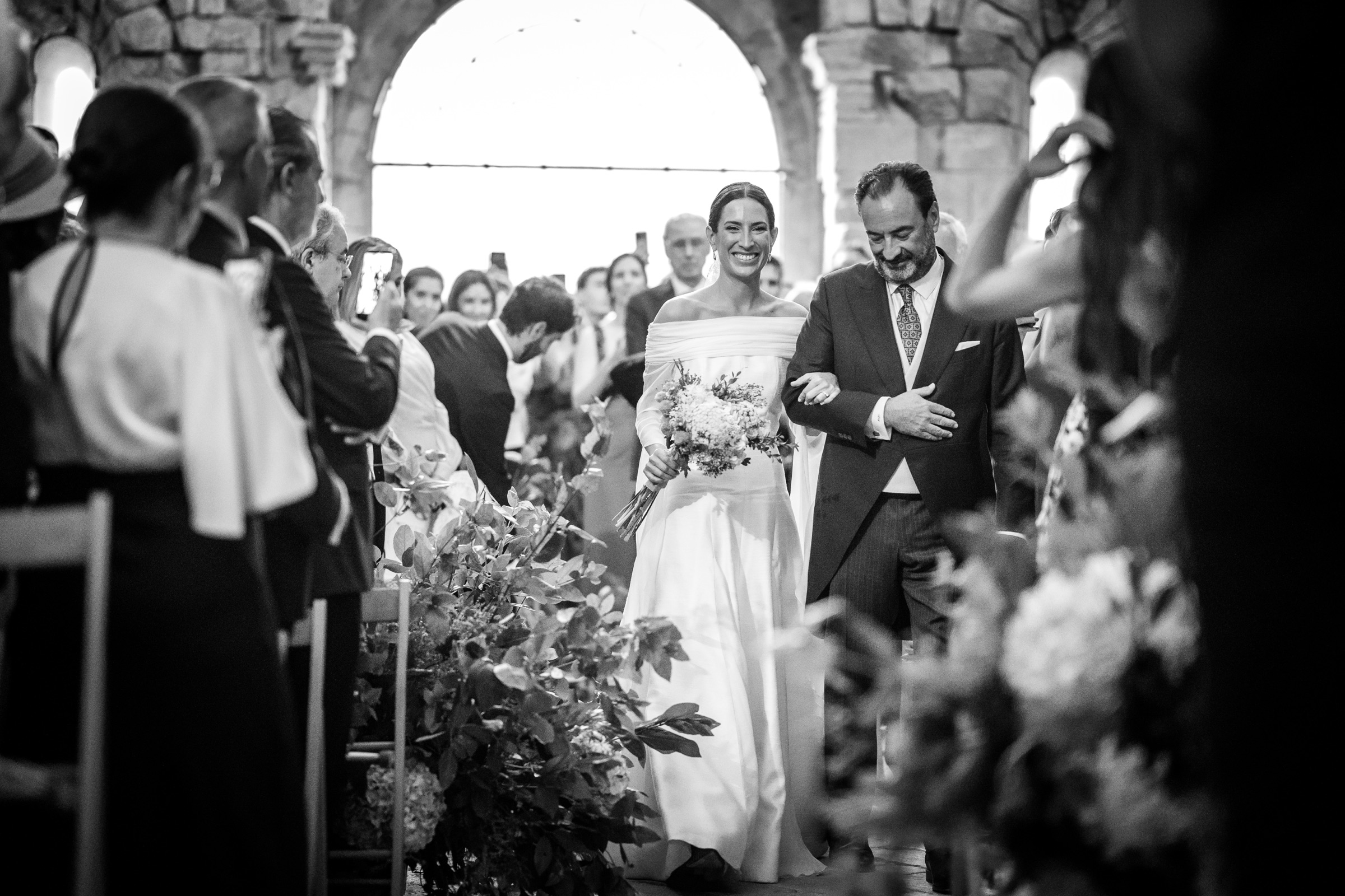 The bride entering on her father's arm beneath the great Romanesque arch of San Juan de la Peña, guests lining the flower-edged aisle