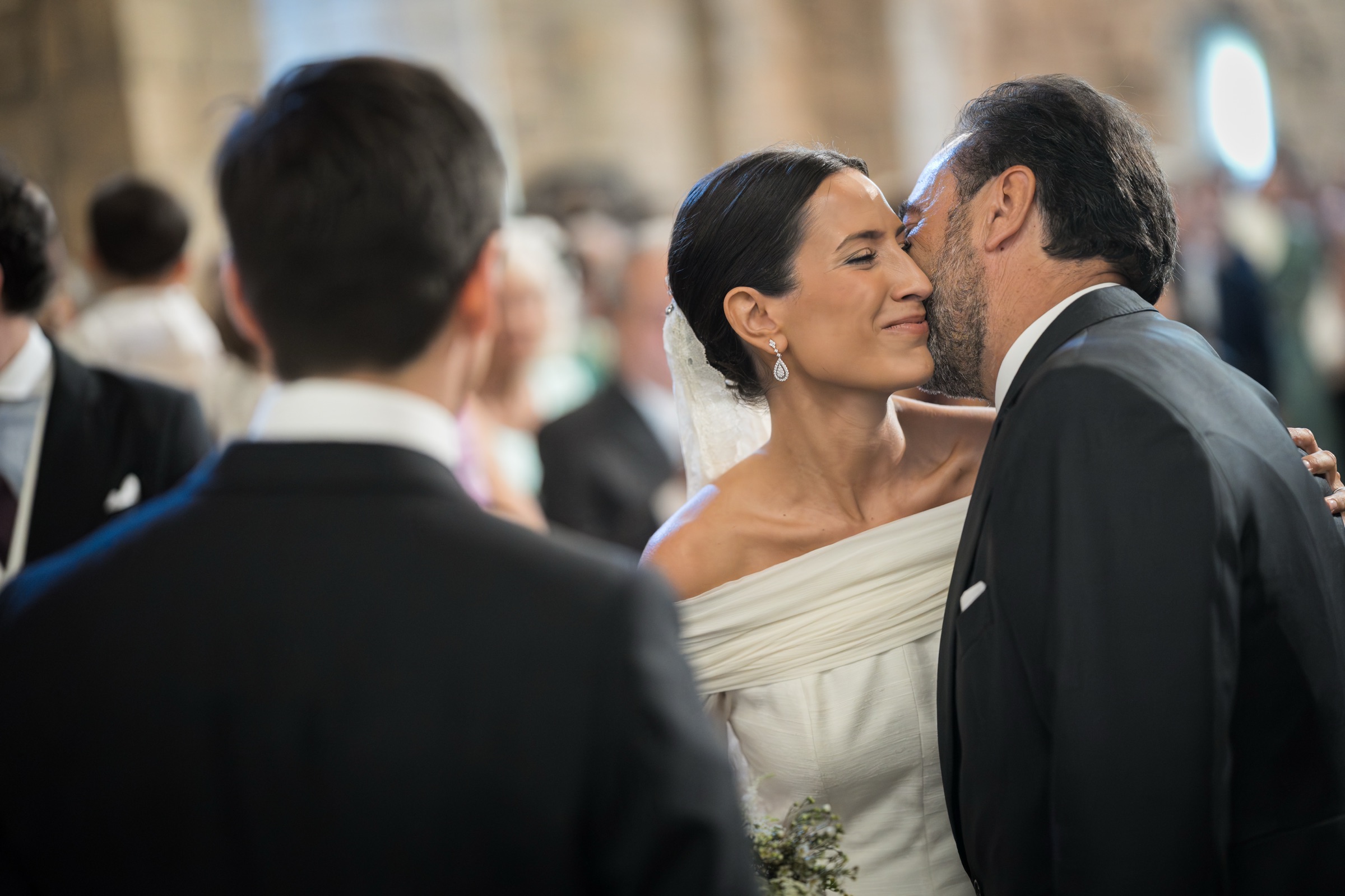 Her father's kiss on the bride's cheek at the moment of handover, the groom's shoulder framing the scene