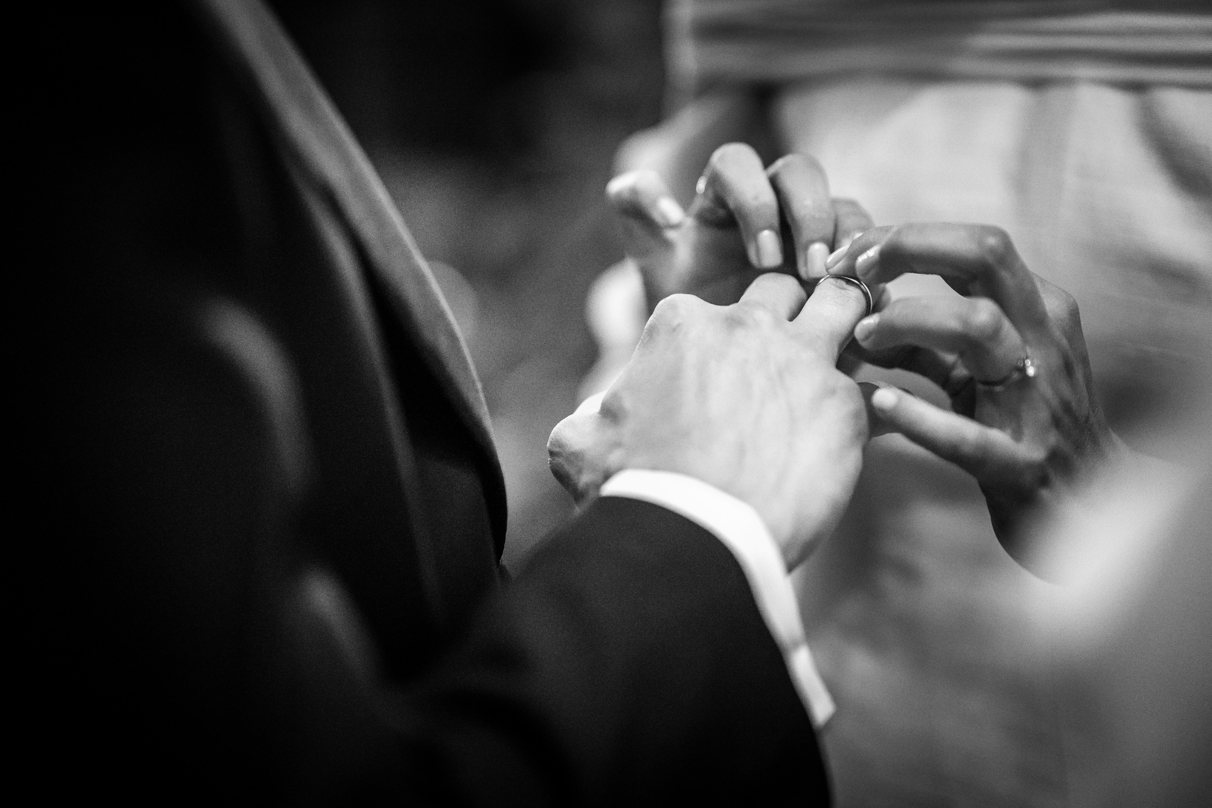 The bride's fingers slide the wedding band onto the groom's hand, white cuff and dark tailoring filling the frame