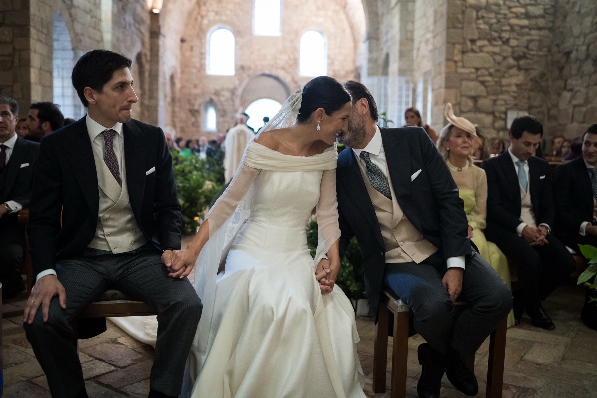 Seated between their witnesses, bride and groom lean forehead to forehead in the bare Romanesque stone nave