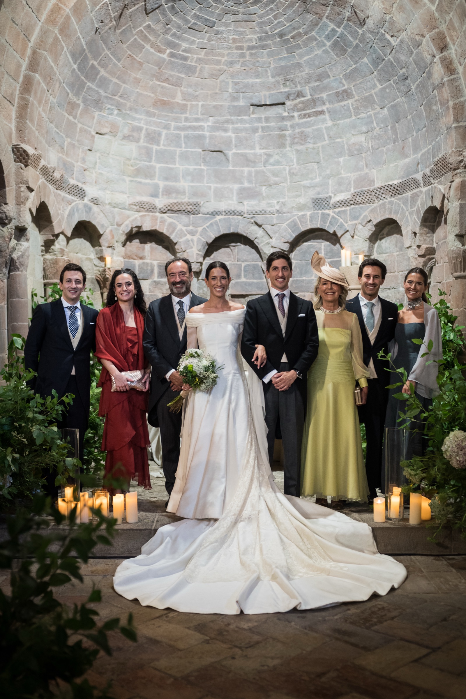 The couple with their parents and siblings beneath the candlelit stone apse, the bride's train fanned across the flagstones