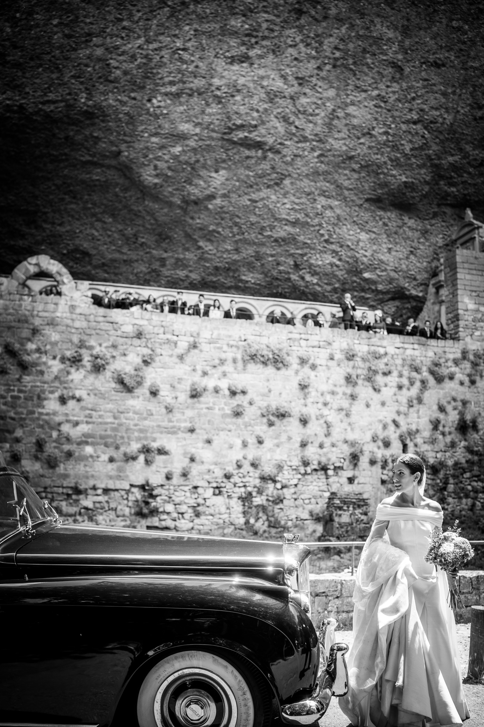 The bride glances back beside a vintage black car beneath the immense rock overhang of San Juan de la Peña