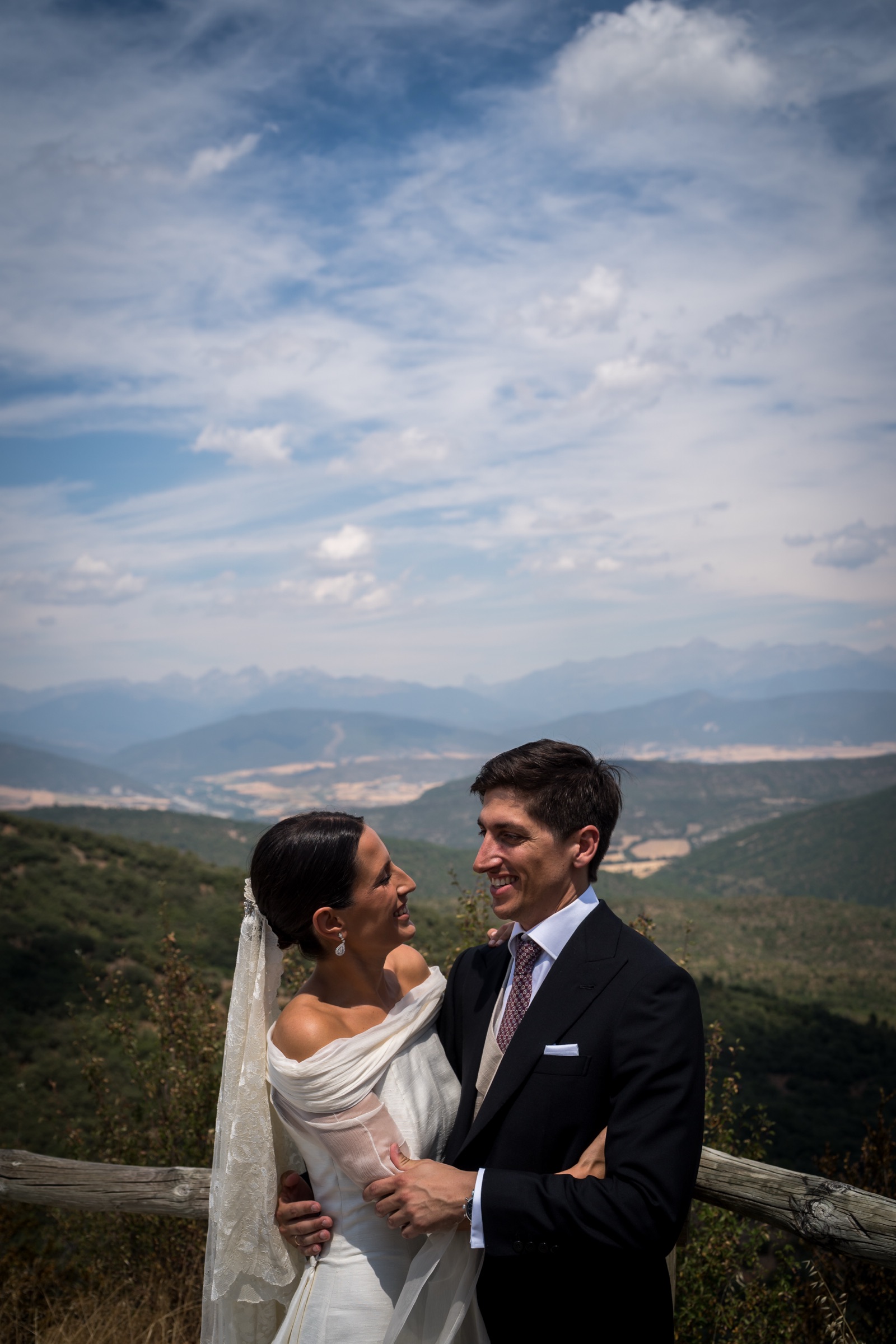 The couple embrace at a rustic wooden fence high above the valley, the Pyrenees fading to the horizon under towering clouds