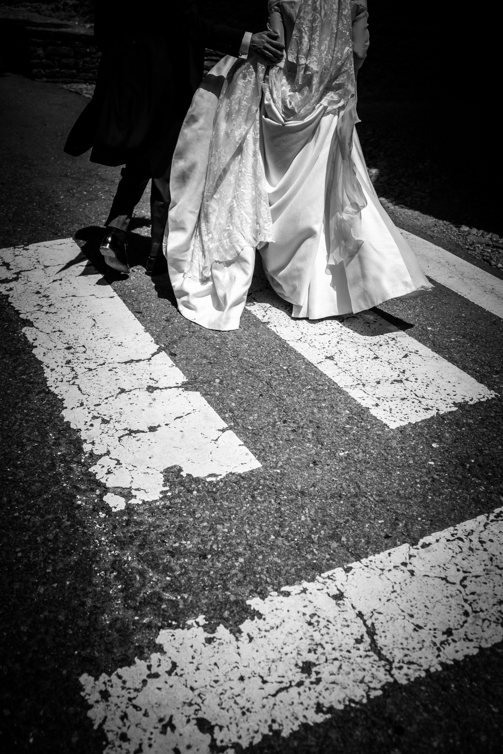 The couple's trailing lace train and polished shoes crossing a sun-bleached zebra crossing, hard shadow slicing the asphalt
