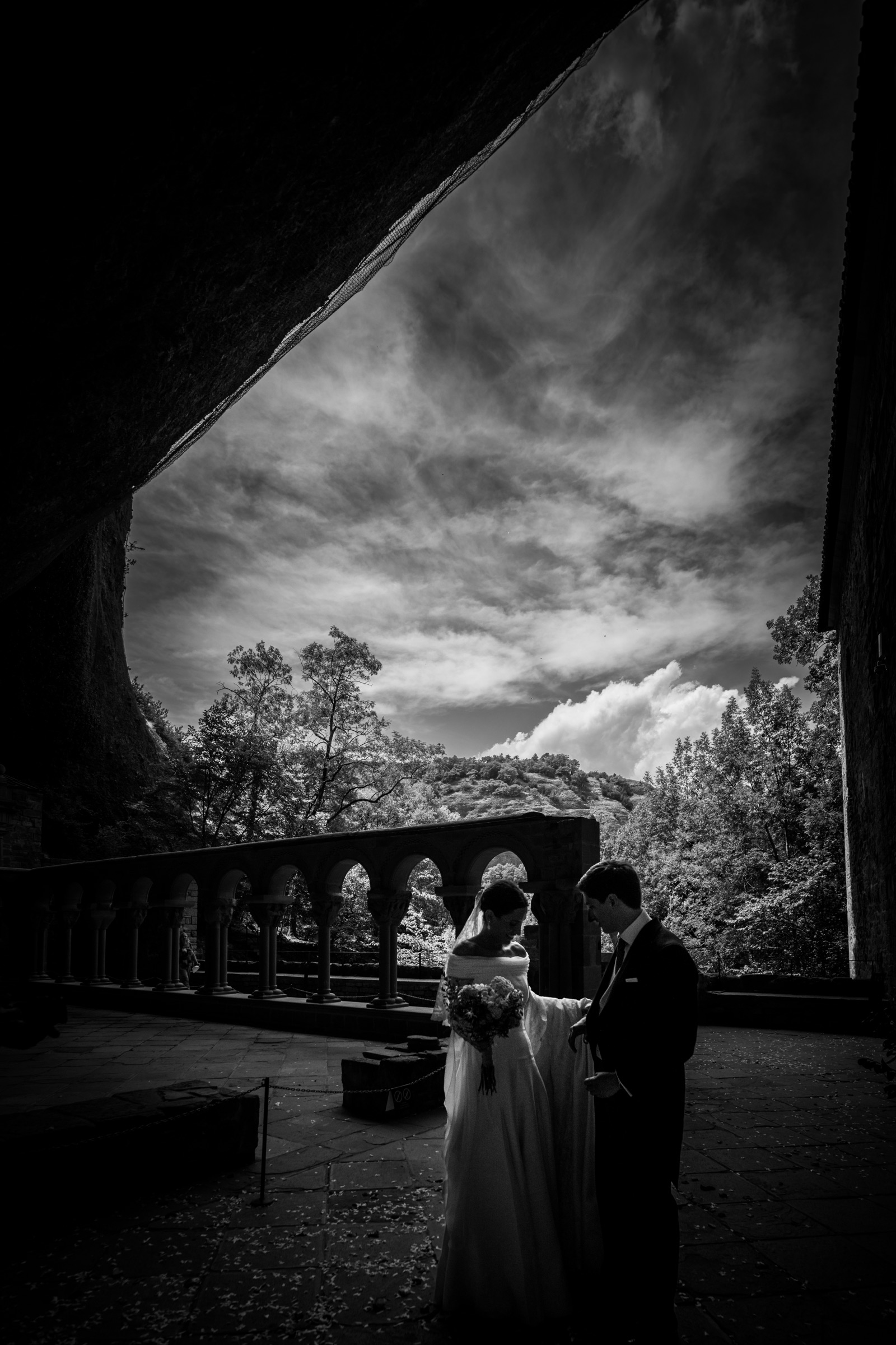 Bride and groom small beneath the great rock overhang of San Juan de la Peña, the Romanesque cloister arcade and a streaked sky opening beyond