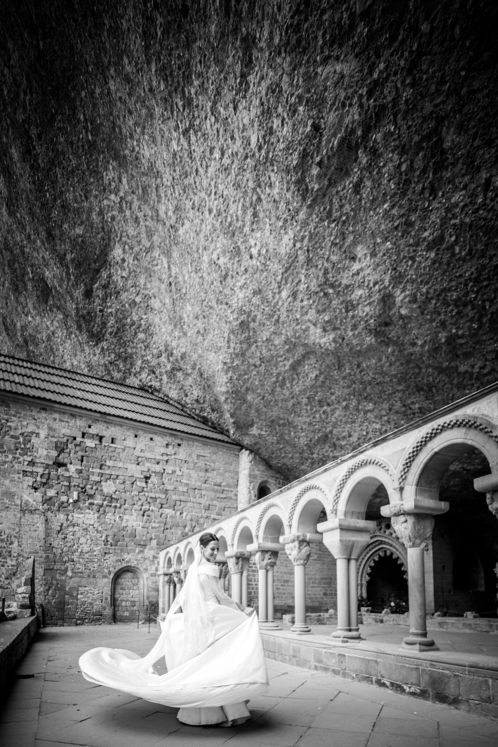 Bride mid-twirl on the cloister terrace, her dress flaring white beneath the cliff face of San Juan de la Peña