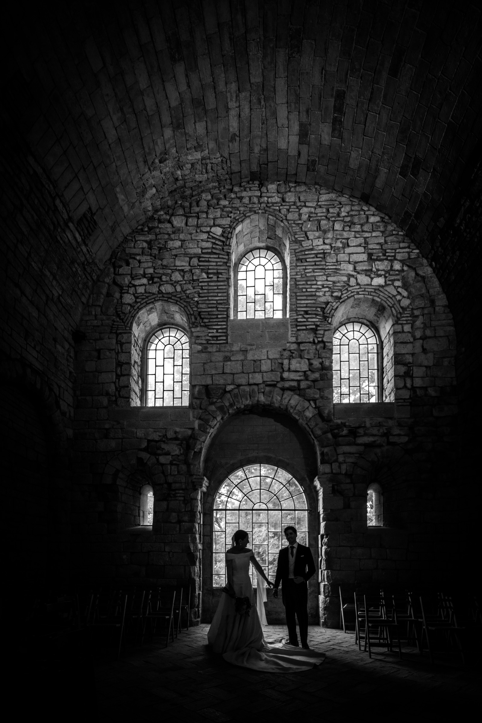 Bride and groom hand in hand beneath the vaulted stone nave, dwarfed by the glowing arched windows of the monastery church