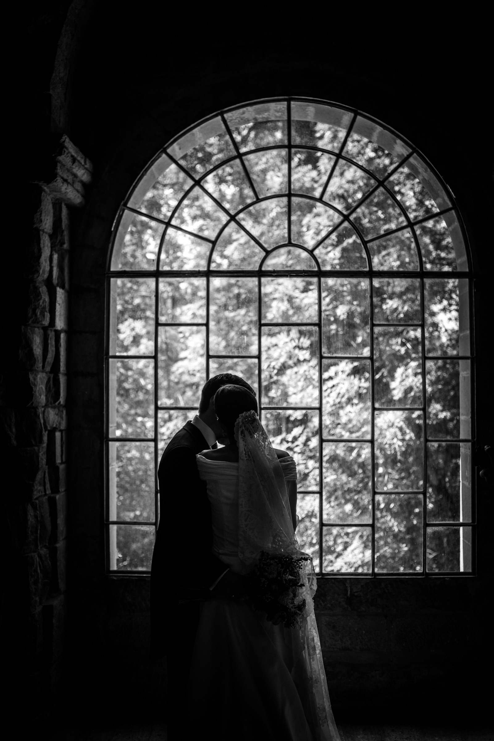 Couple kissing in near-silhouette before a great leaded arch window, her embroidered veil catching the light of the trees beyond the glass