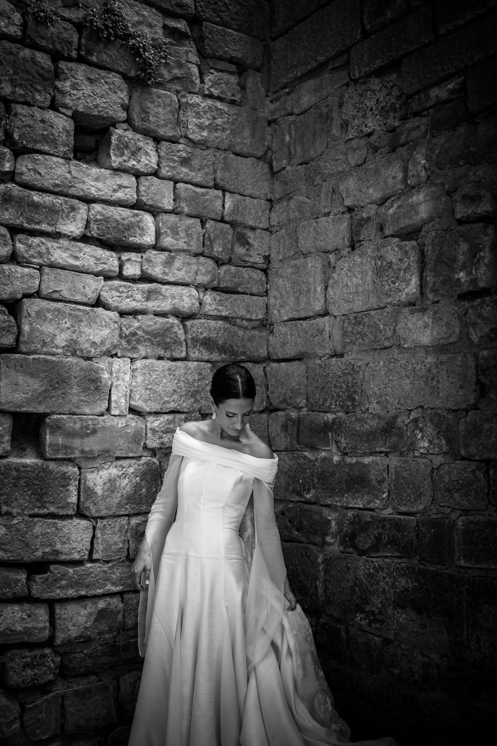 Bride looking down and gathering her skirt, her gown luminous against the dark rubble-stone corner of the monastery wall