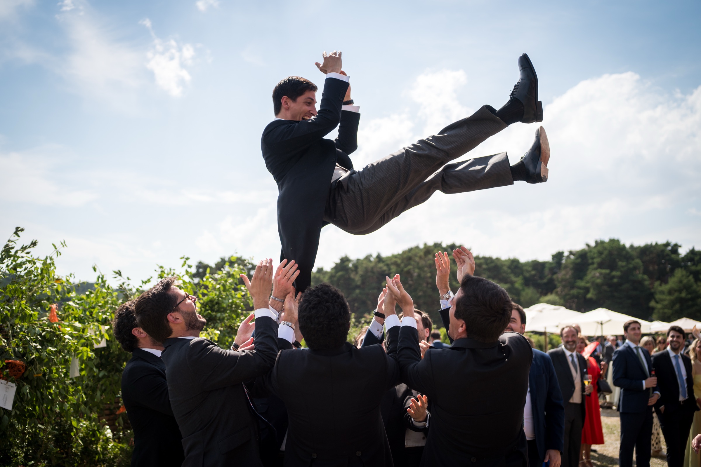 Groom tossed high above his friends' raised hands against a blue Aragonese sky, parasols and cocktail guests scattered below
