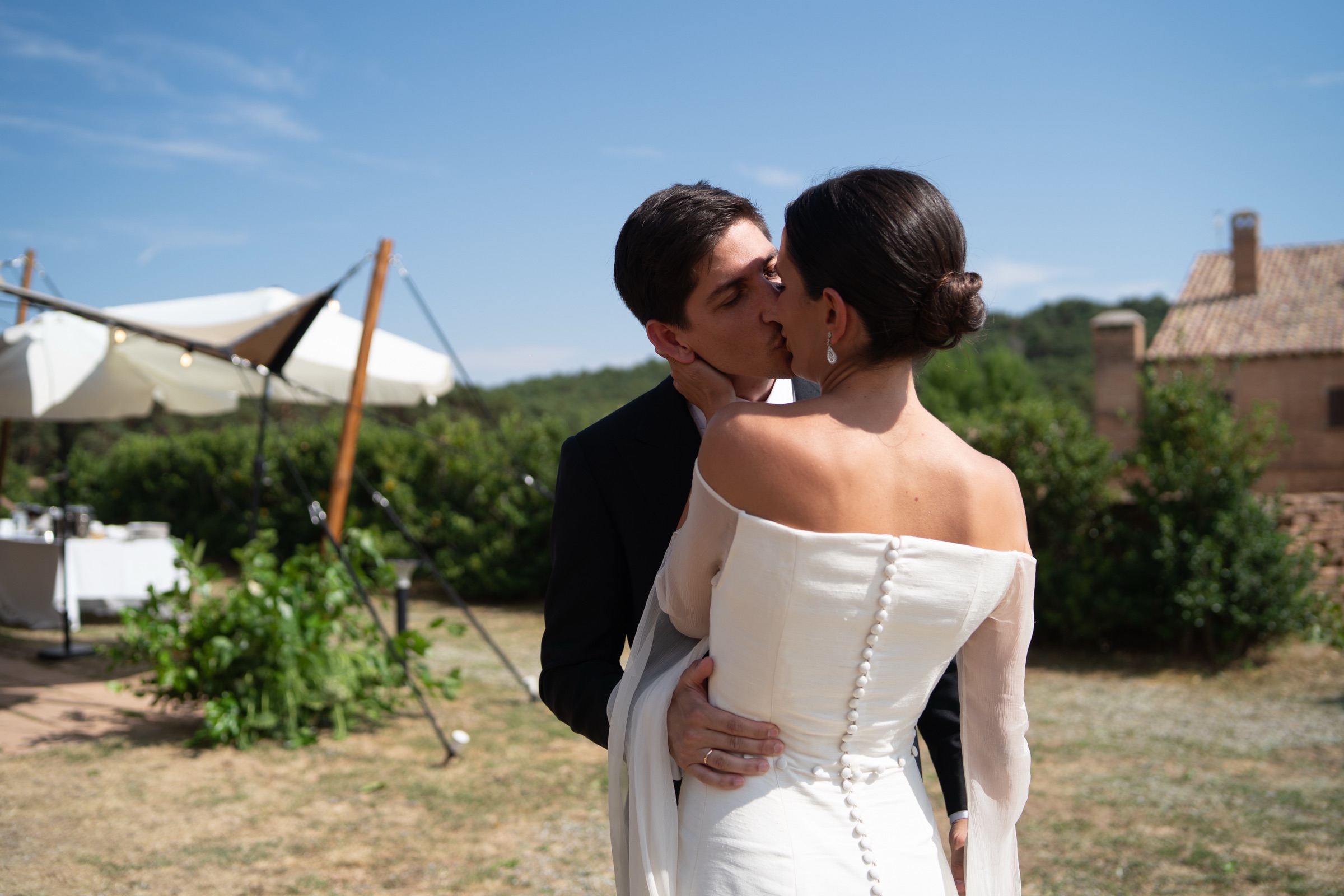 Bride and groom kissing beside the cocktail marquee, the buttoned silk back of her dress in full sun