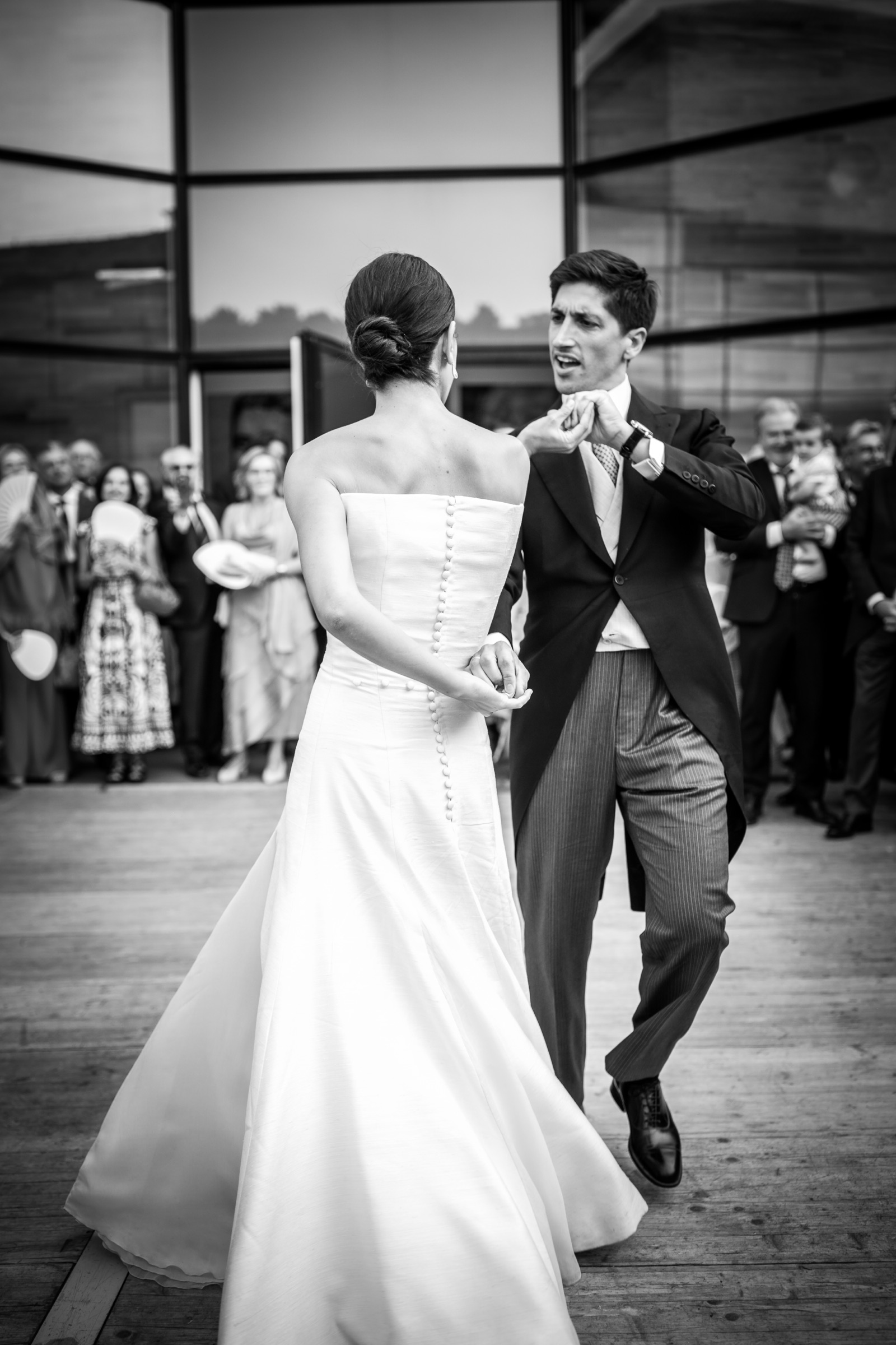 First dance seen over the bride's bare shoulders, the groom singing along as guests watch from the glass wall of the pavilion