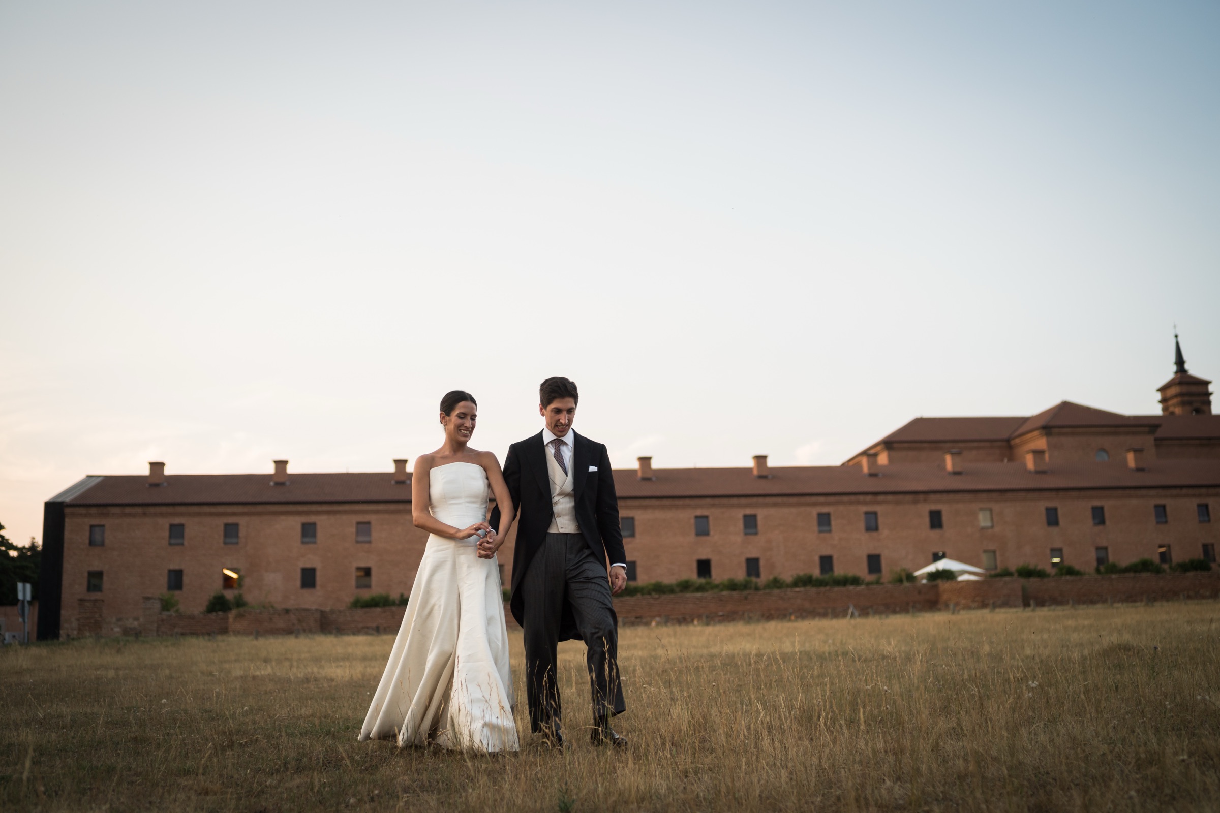 Bride and groom crossing a dry summer meadow at dusk, the brick facade of the new monastery behind them