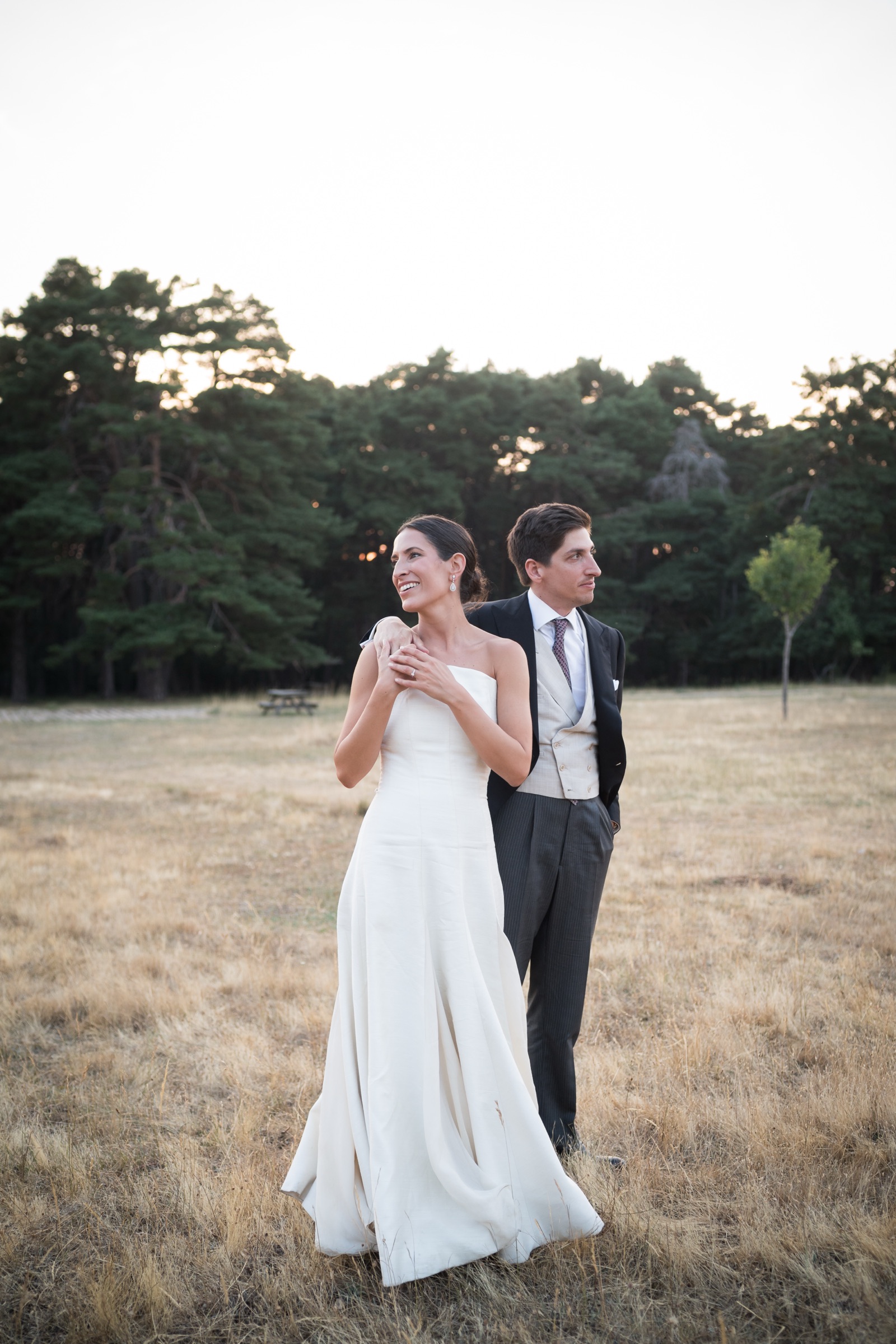 Bride and groom back to back in a dry golden meadow at dusk, each gazing a different way, the pine forest dark behind them