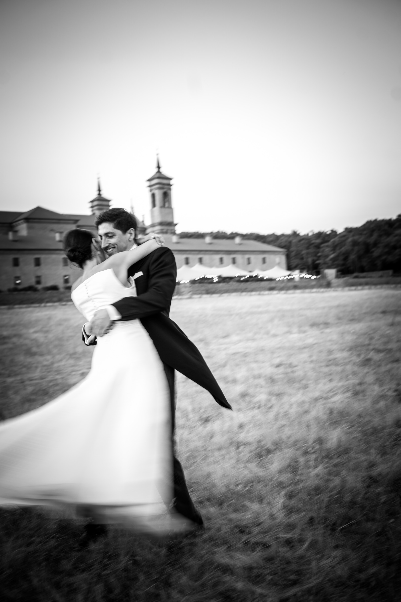 The couple mid-spin in the dry grass, her skirt blurring into motion, the lit reception marquee strung along the monastery wall