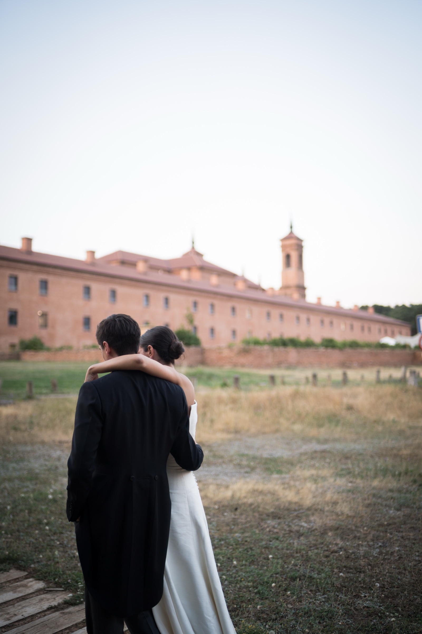 Seen from behind, the couple with arms around each other facing the long brick facade and tower of San Juan de la Peña in the last evening light